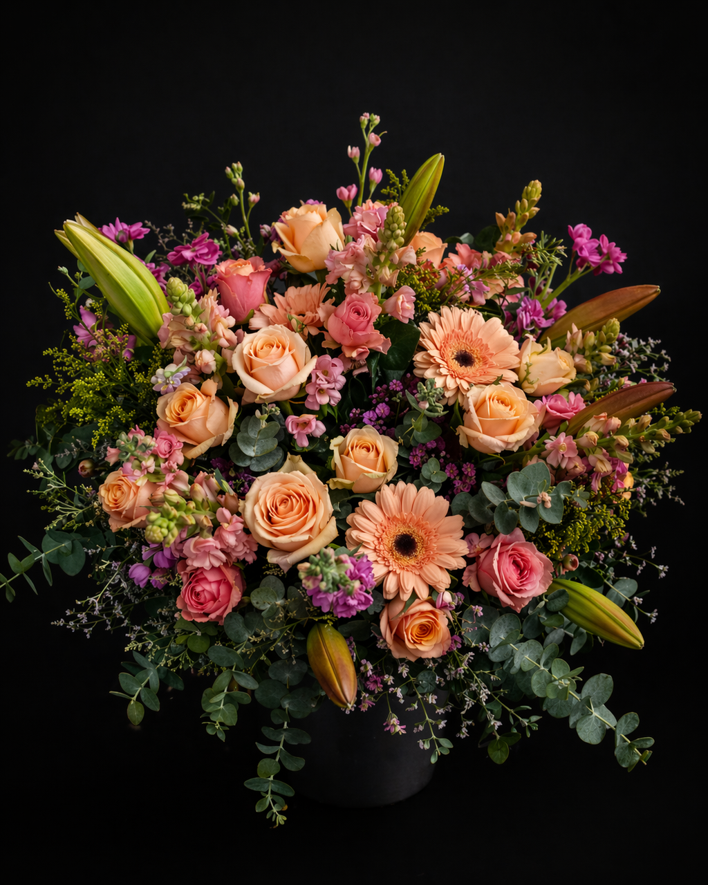 Bouquet with peach roses, pink stargazer lilies, peach gerbera daisies, pink roses, pink snapdragons, pink stocks, yellow golden rod, pink limonium and grey pennygum on a black background.