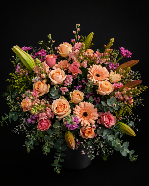 Bouquet with peach roses, pink stargazer lilies, peach gerbera daisies, pink roses, pink snapdragons, pink stocks, yellow golden rod, pink limonium and grey pennygum on a black background.