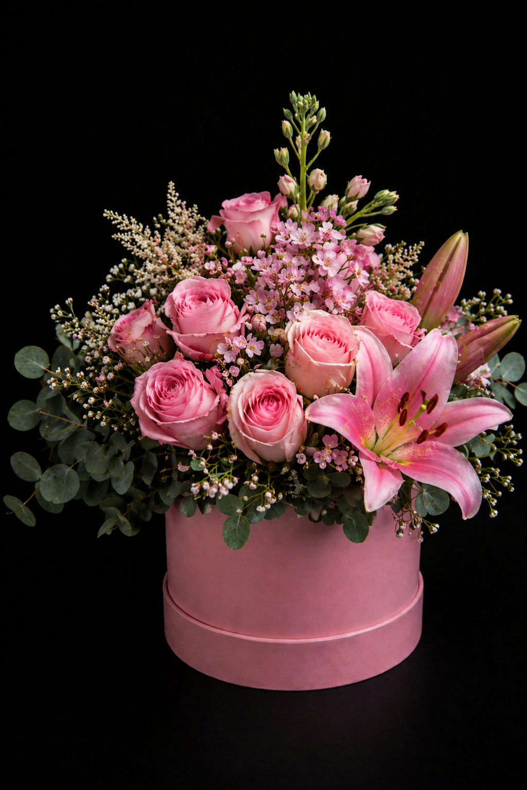 Pink posy style flower arrangement, with pink roses, pink stargazer lily, pink stocks, pink wax and grey pennygum in a pink medium sized hatbox on a black background.